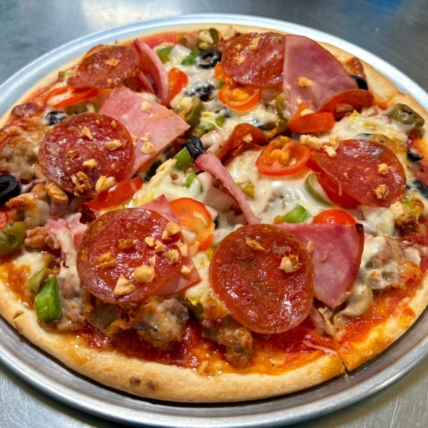Whole supreme pizza with pepperoni, ham, olives, peppers, tomatoes, and cheese on a metal pan.