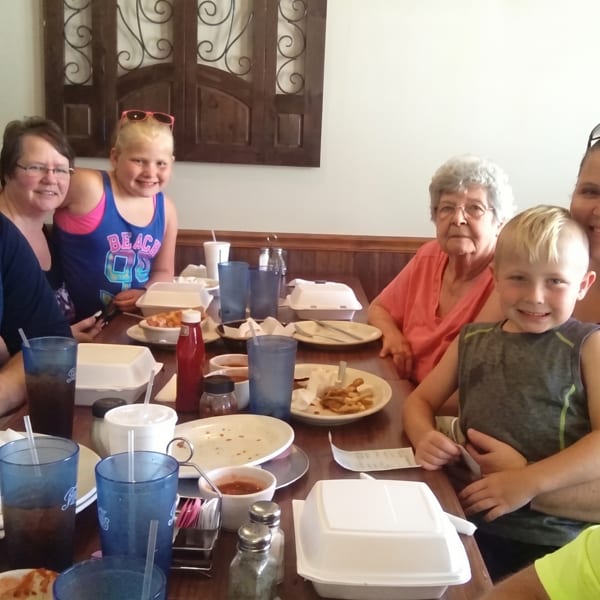 A large family group, including children and seniors, smiling at a restaurant table with plates and drinks.